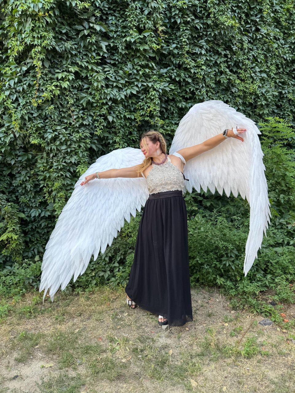 May include: A person wearing large white angel wings, a black skirt, and a silver top, standing in front of a green leafy background. The wings are large and feathered, with a wide wingspan. The person is smiling and has their arms outstretched.