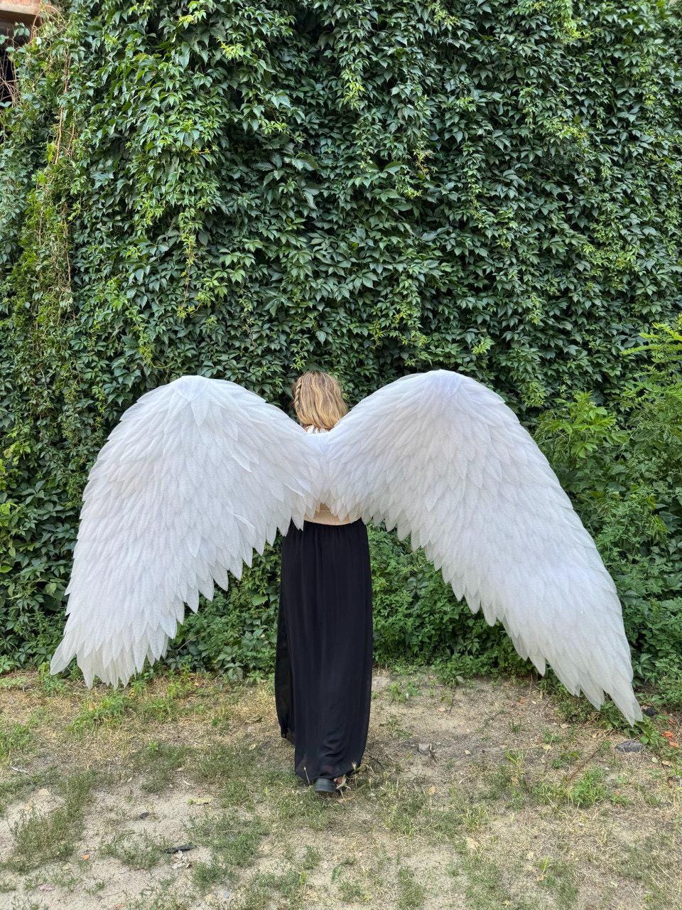May include: A person wearing large, white angel wings against a backdrop of green foliage. The wings are made of layered, feathery material, and the person is wearing a long, black dress. The wingspan appears to be several feet wide.