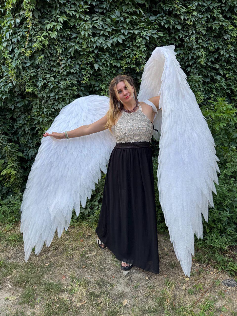 May include: A person wearing large, white feathered angel wings, a beaded top, and a long black skirt. The wings are spread wide, and the person is standing in front of a green leafy background.