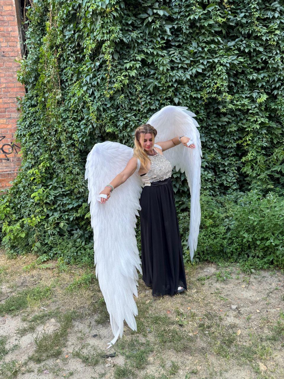 May include: A person wearing large, white angel wings, standing in front of a wall covered in green vines. The wings are detailed with individual feathers. The person is wearing a black skirt and a silver top.