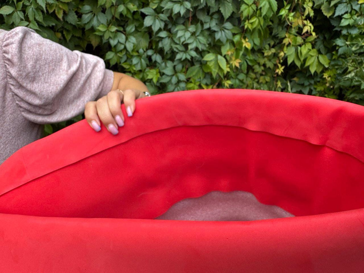 May include: A vibrant red, curved object, possibly a piece of furniture or decor, with a soft, velvety texture. The object is partially visible, with a person's hand resting on the edge. The background features lush green foliage.