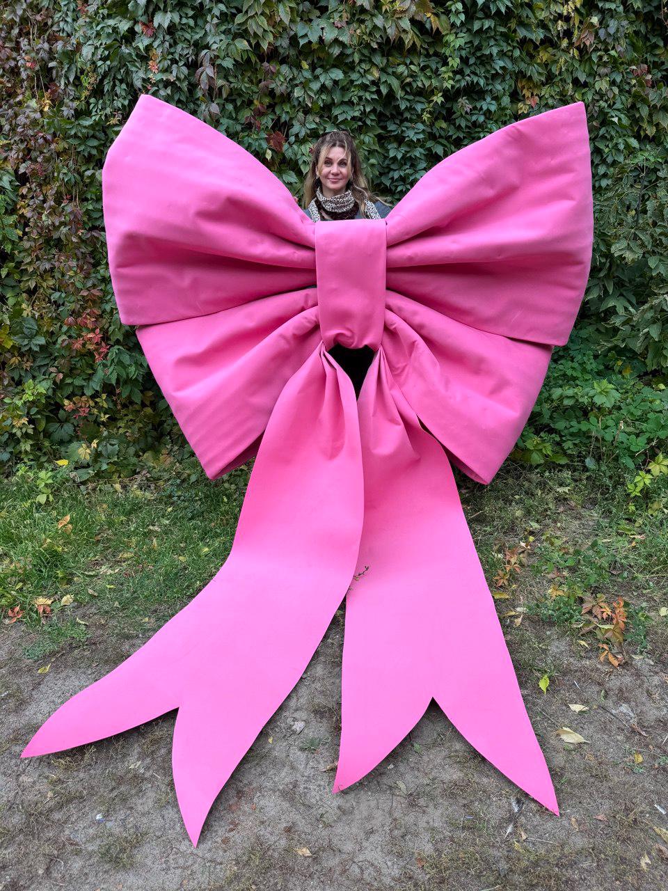 May include: A large, bright pink bow with long, pointed tails. The bow is made of a smooth fabric and is positioned in front of a green leafy background. The bow's size suggests it could be used for a decorative display or event.