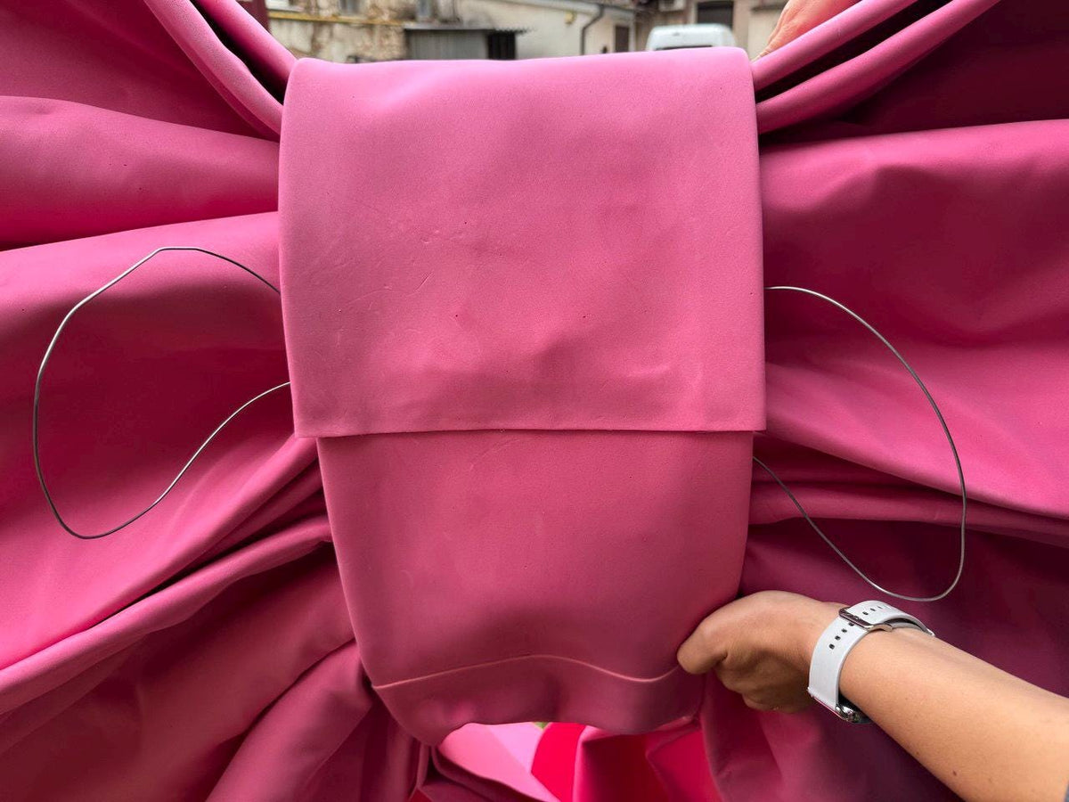May include: A large, vibrant pink bow made of fabric. The bow has a rectangular center and flowing, folded fabric on either side. Thin, silver wire accents the bow's shape. The bow is held by a person wearing a white watch.