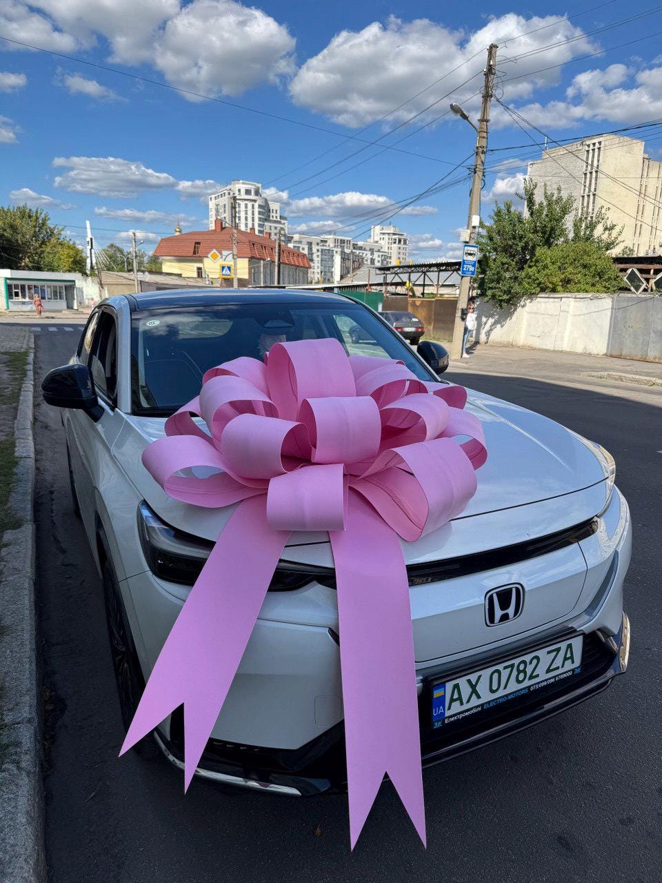 May include: A white car with a large pink bow on the hood, parked on a city street. The bow is made of wide, pink ribbons. The car has a black roof and a license plate.