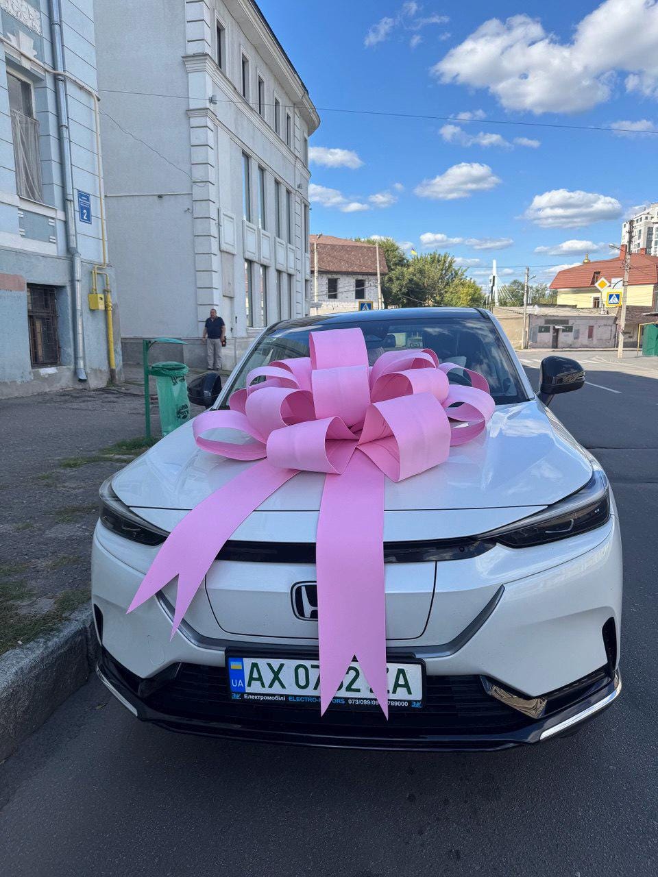 May include: A white car with a large pink bow on the hood. The car has a black front bumper and headlights. The license plate reads "AX 07 2 A". The car is parked on a street with buildings in the background.