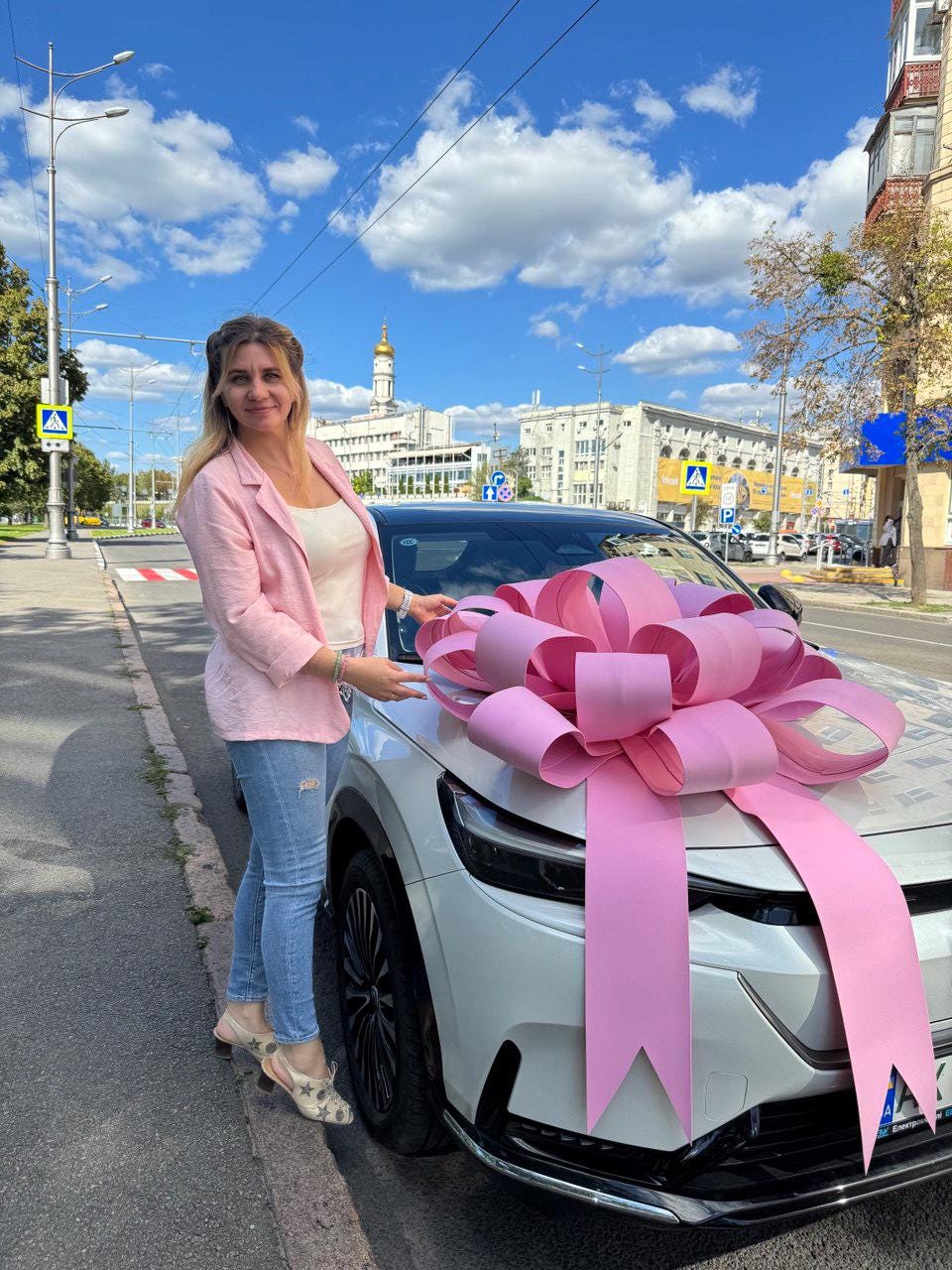 May include: A white car with a large pink bow on the hood. The bow is made of wide, looped ribbons. The car is parked on a city street with a woman standing next to it. The woman is wearing a pink blazer and blue jeans.