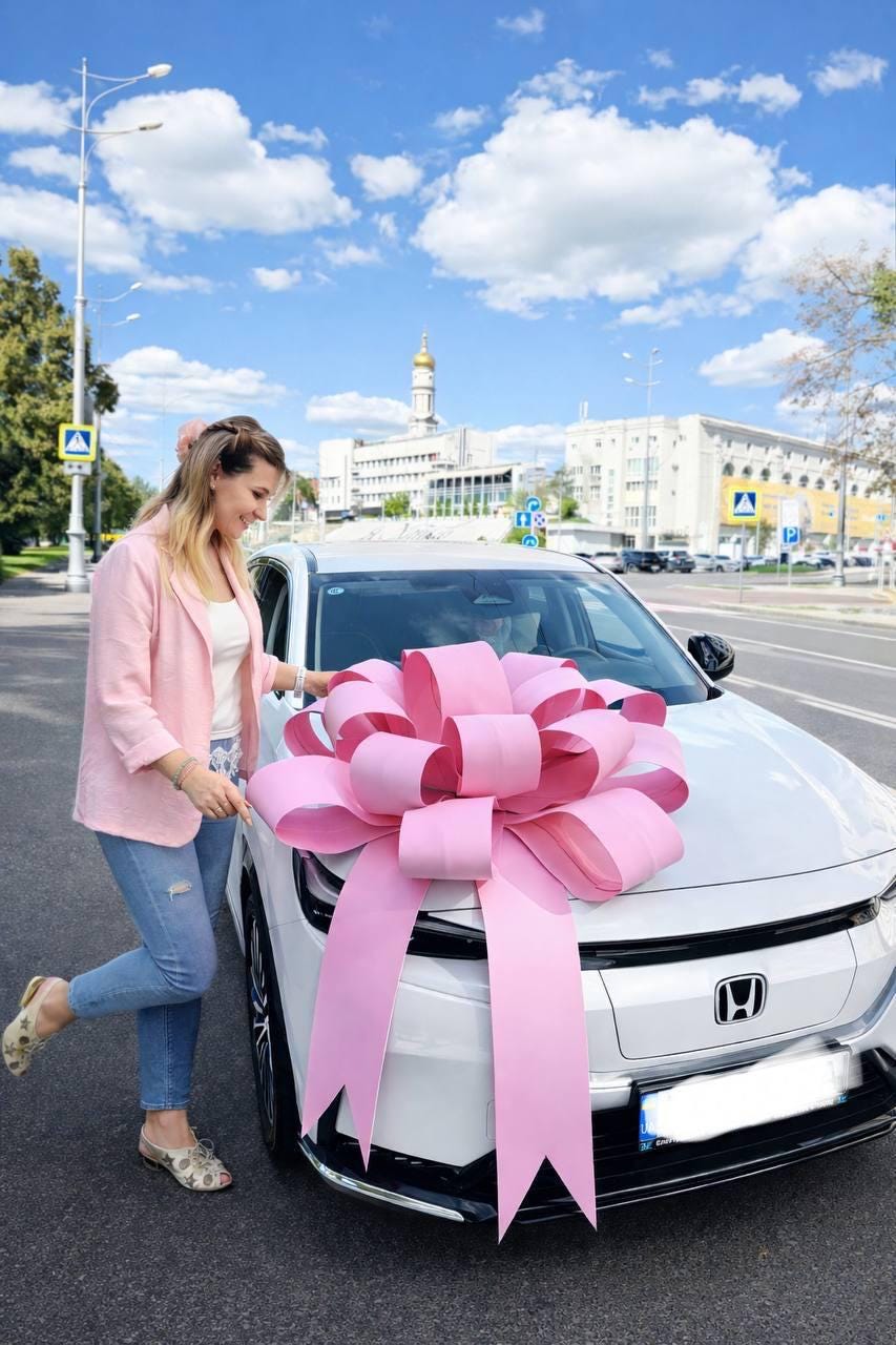 May include: A white car with a large pink bow on the hood. The car is parked on a city street with a woman standing next to it. The woman is wearing a pink blazer, white top, and blue jeans.