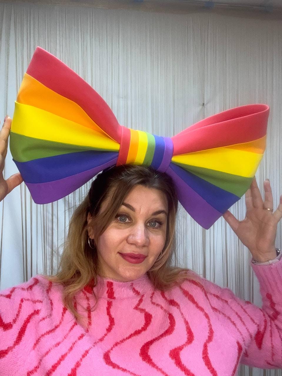 May include: A large, colorful rainbow bow, featuring red, orange, yellow, green, blue, and purple stripes. The bow is worn on the head. The person is wearing a pink sweater with red wavy stripes.