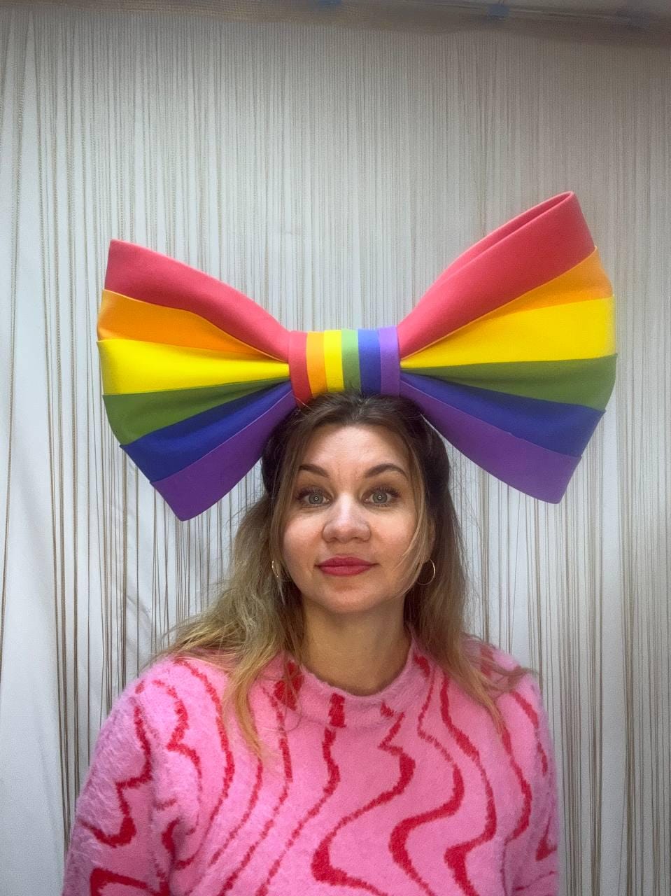 May include: A large, rainbow-colored bow headband. The bow features the colors of the Pride flag: red, orange, yellow, green, blue, and purple. The bow is worn on the head. The person is wearing a pink sweater.