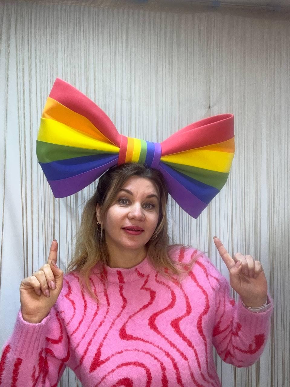 May include: A large, colorful rainbow bow, worn on the head. The bow features the colors of the rainbow: red, orange, yellow, green, blue, and purple. The person is wearing a pink sweater with a wavy red pattern.