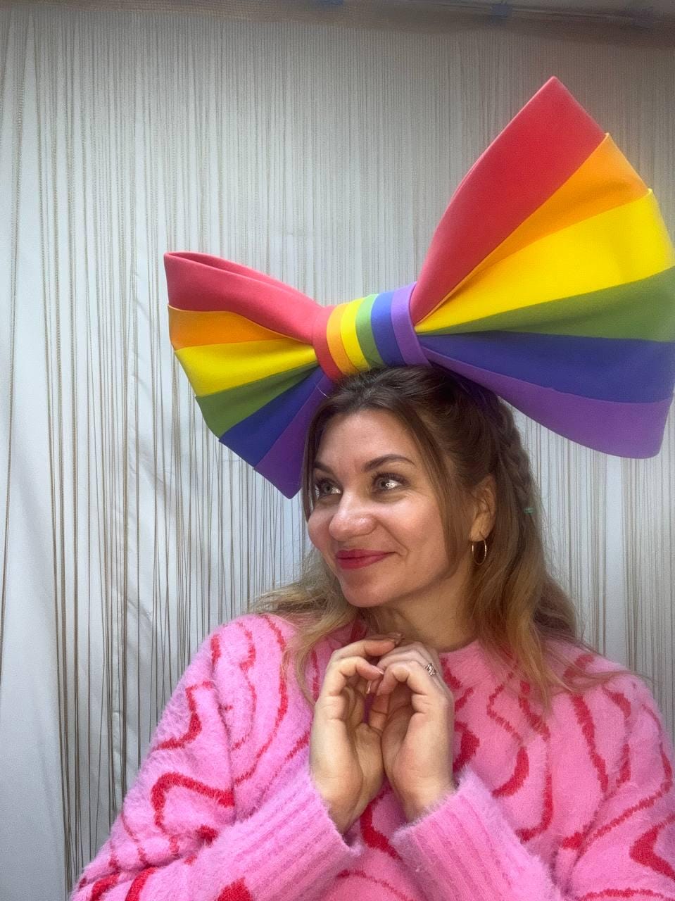 May include: A large, rainbow-colored bow, with red, orange, yellow, green, blue, and purple stripes, is worn on the head. The person is wearing a pink sweater with a red pattern.