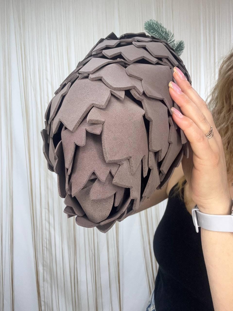 May include: A large, brown, decorative object resembling a pinecone. The object is made of overlapping, petal-shaped pieces. A small sprig of green foliage is attached to the top. The object is held by a person.