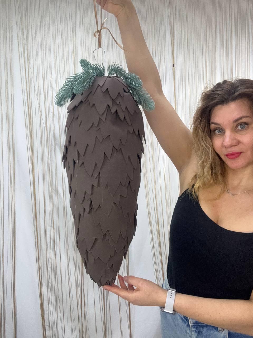 May include: A large, brown, cone-shaped decoration with a textured surface, resembling a pine cone. The top is adorned with faux green pine needles. It is suspended by a thin, brown ribbon.