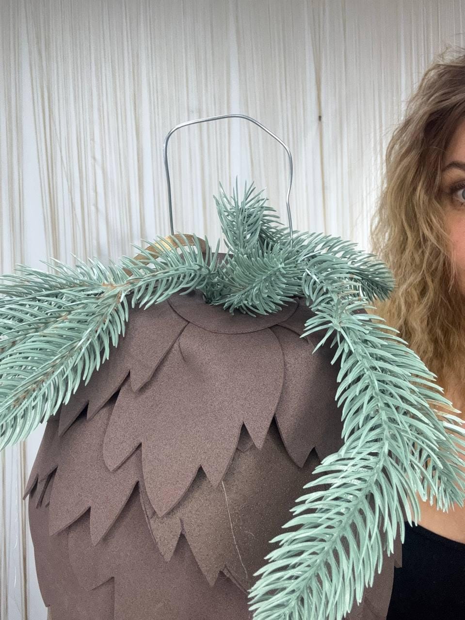 May include: A brown, cone-shaped decorative item with a silver handle and faux evergreen branches. The cone is made of overlapping brown petal-shaped pieces. The evergreen branches are a muted green color.