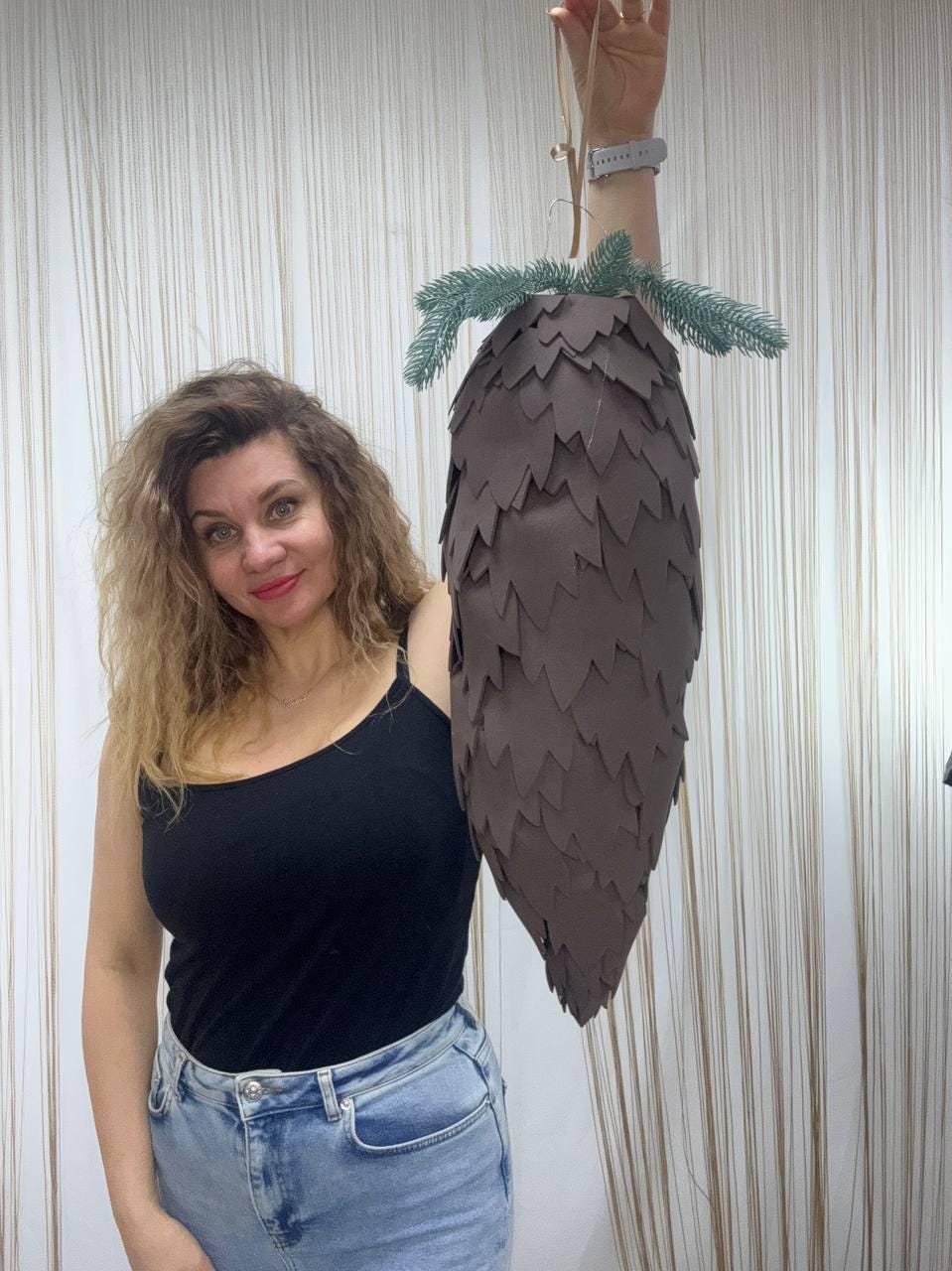 May include: A large, brown, cone-shaped decorative item with a textured surface, resembling a pine cone. The top features green, artificial pine needles. It is suspended by a thin, beige string. The item is held up by a person.