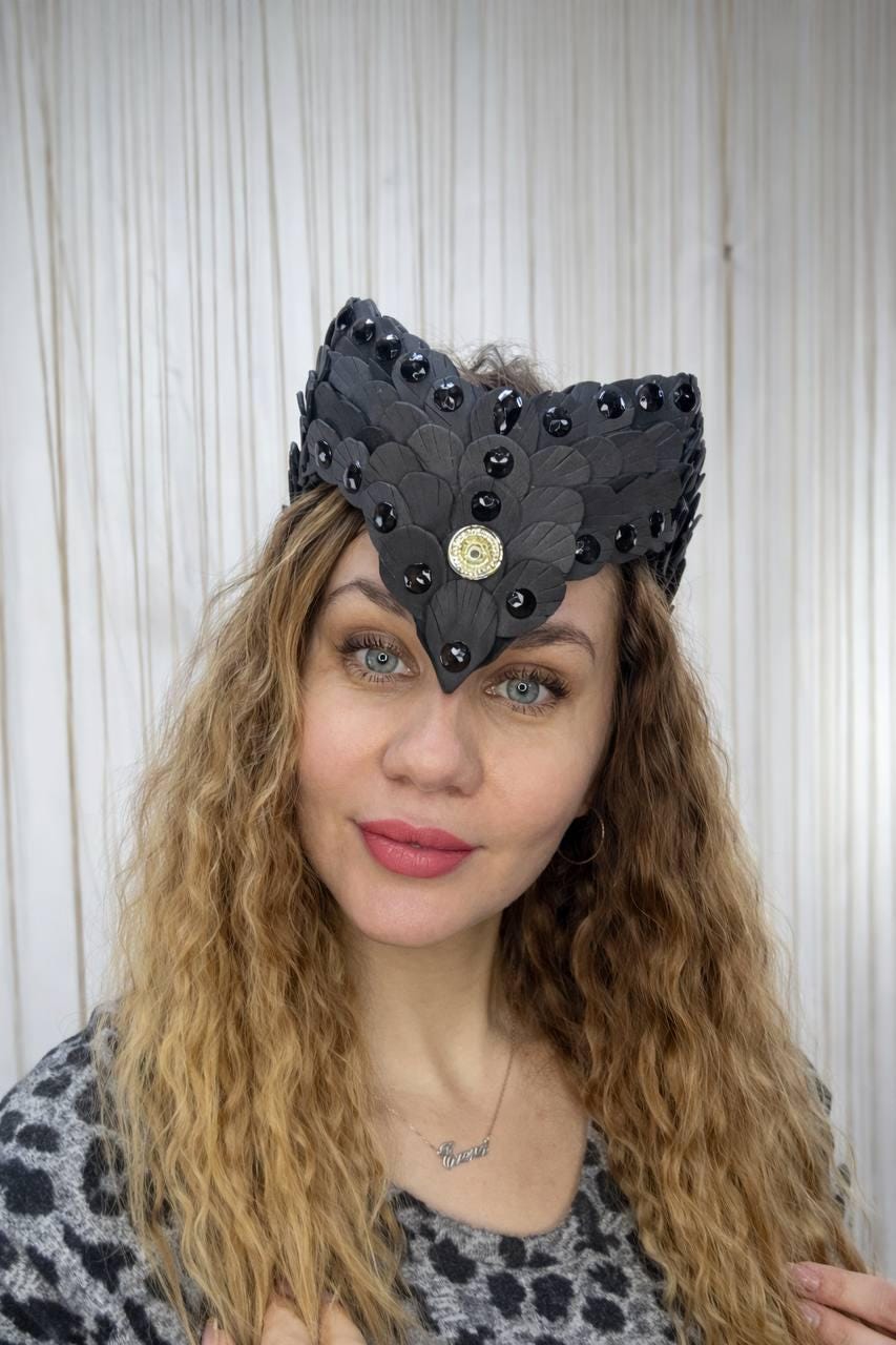 May include: A black headpiece with a feather design and black gemstones. The headpiece has a gold-colored circular embellishment in the center. The woman in the image has long, wavy blonde hair and is wearing a gray and black patterned top.