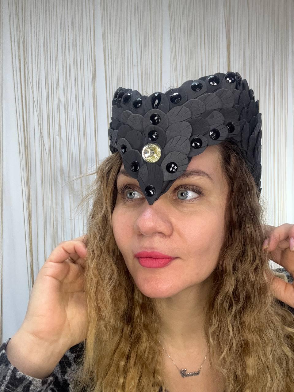 May include: A black headpiece with a layered, feather-like design, adorned with black and gold-colored embellishments. The headpiece has a central, pointed design and is worn on the head. The headpiece is a unique accessory.