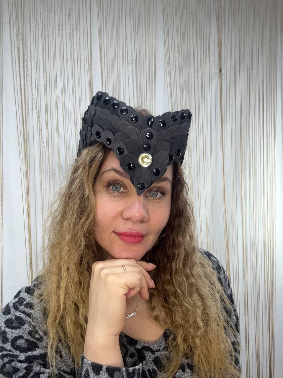 May include: A black headpiece with a layered, scale-like design, adorned with black gems and a central gold-colored emblem. The headpiece is worn by a person with long, wavy blonde hair and a patterned top.