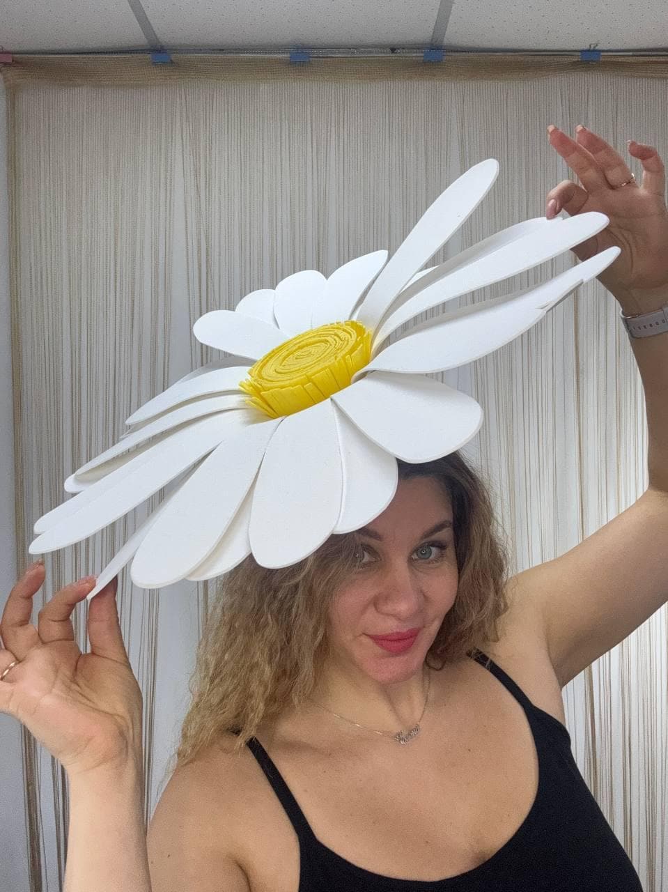 May include: A large, white daisy flower hat with a bright yellow center. The hat has oversized petals and is worn on the head. The hat is a whimsical accessory.