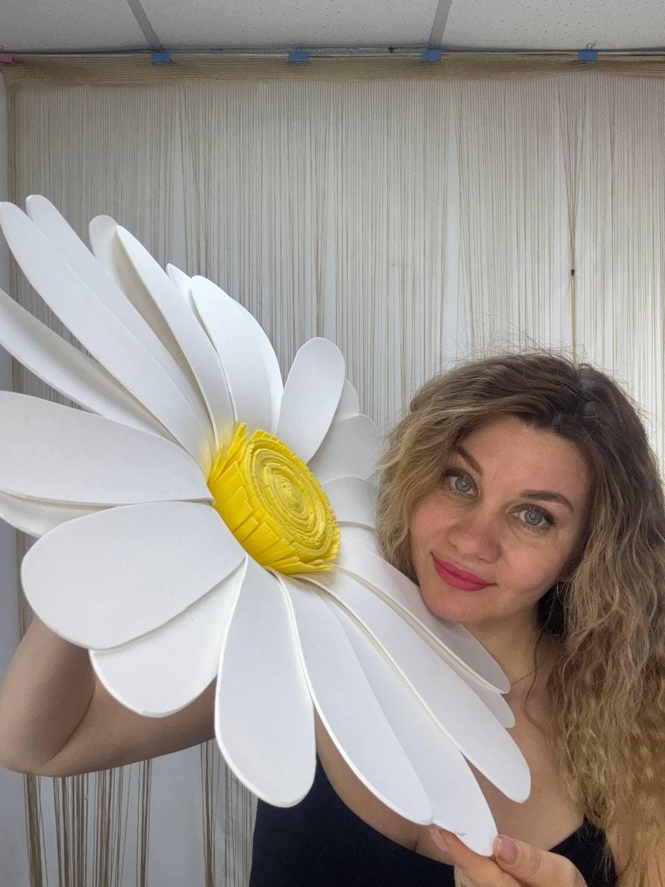 May include: A large, white daisy flower with a yellow center. The flower is held up in front of a person. The petals are large and layered, and the center is a textured yellow. The person is wearing a black top.
