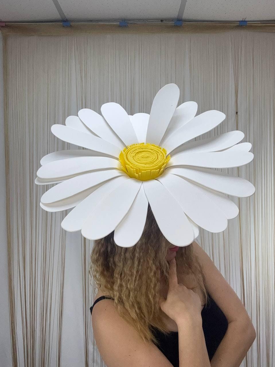 May include: A large, white daisy flower headpiece with a yellow center. The petals are layered and curved, creating a three-dimensional effect. The headpiece is worn by a person wearing a black top.