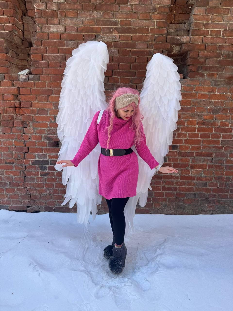 May include: A person wearing a pink dress with a black belt, black leggings, and gray boots with fur trim, posing in front of large white angel wings. The background features a brick wall and snow.