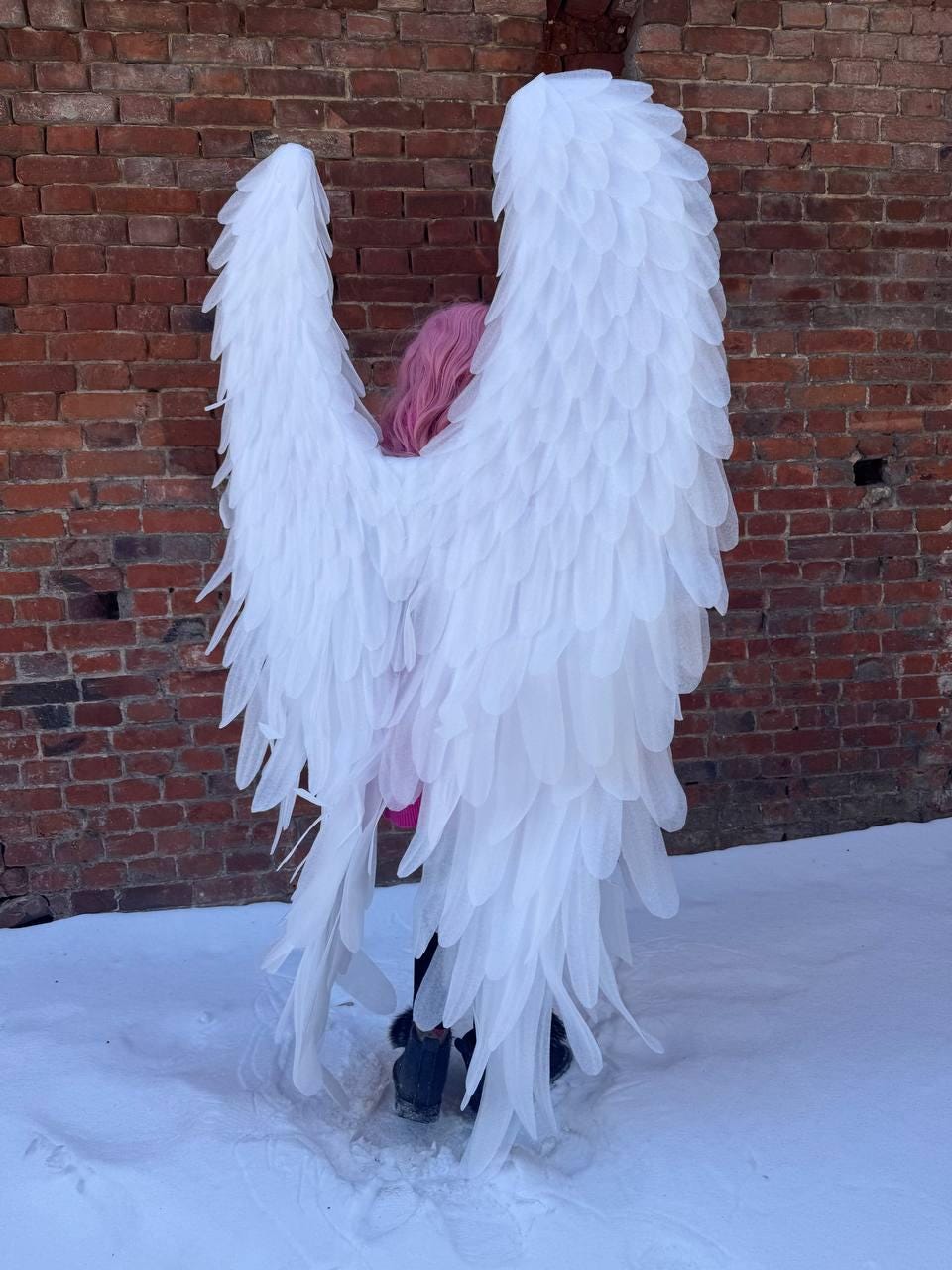May include: Large, white angel wings with layered feather-like details. The wings are worn by a person with pink hair, standing in front of a brick wall and snow. The wings are a costume accessory.