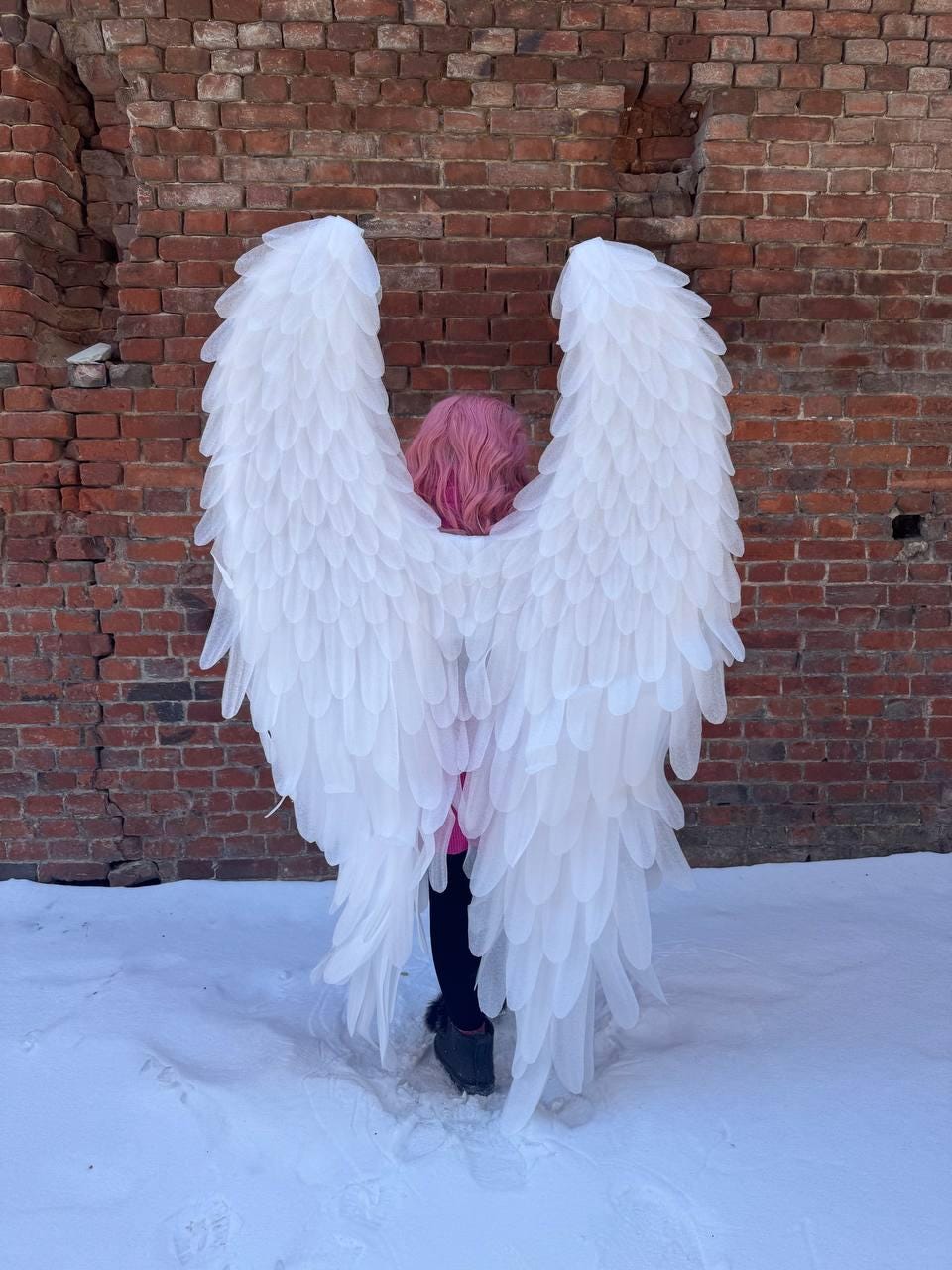 May include: Large, white angel wings with a layered feather design. The wings are spread wide, covering the back of a person with pink hair. The background is a weathered brick wall and snow.