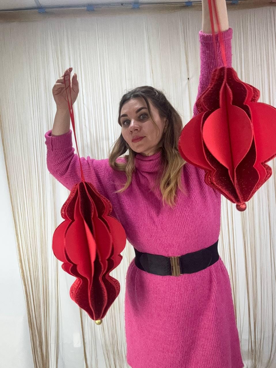 May include: Two red, paper lantern-style ornaments with a layered design are suspended by red ribbons. The ornaments have a textured, glittery appearance and a gold-colored bead at the bottom. The background is a light-colored curtain.