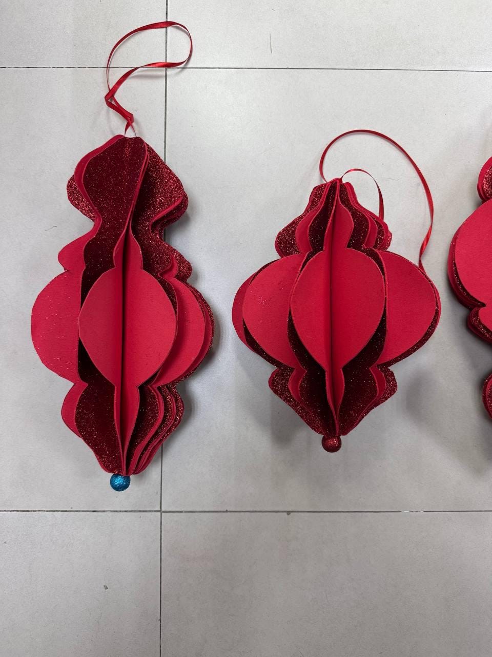 May include: Red paper ornaments with a layered design and glitter accents. Each ornament has a ribbon for hanging and a small blue or red bead at the bottom. These festive decorations are perfect for the holiday season.