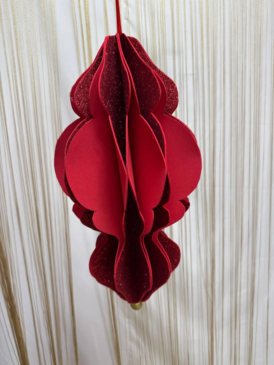 May include: A red, layered paper ornament with a glittery finish. The ornament has a bulbous shape with a scalloped edge design. It is suspended by a red ribbon and has a small gold-colored bead at the bottom.