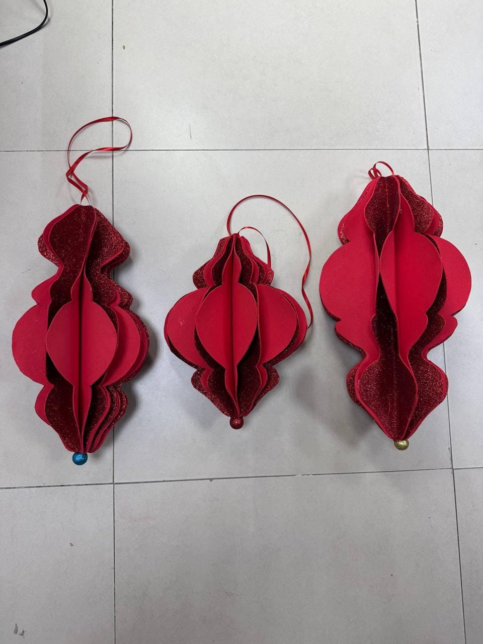 May include: Three red paper ornaments with glitter accents and hanging loops. Each ornament has a unique shape, with a blue or gold bead at the bottom. These decorative ornaments are perfect for holiday or festive decor.