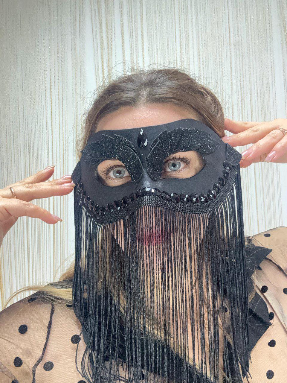 May include: Black masquerade mask with a beaded trim and fringe. The mask features a central jewel and decorative accents. The fringe hangs down, partially obscuring the face. The mask is likely for a costume or special event.