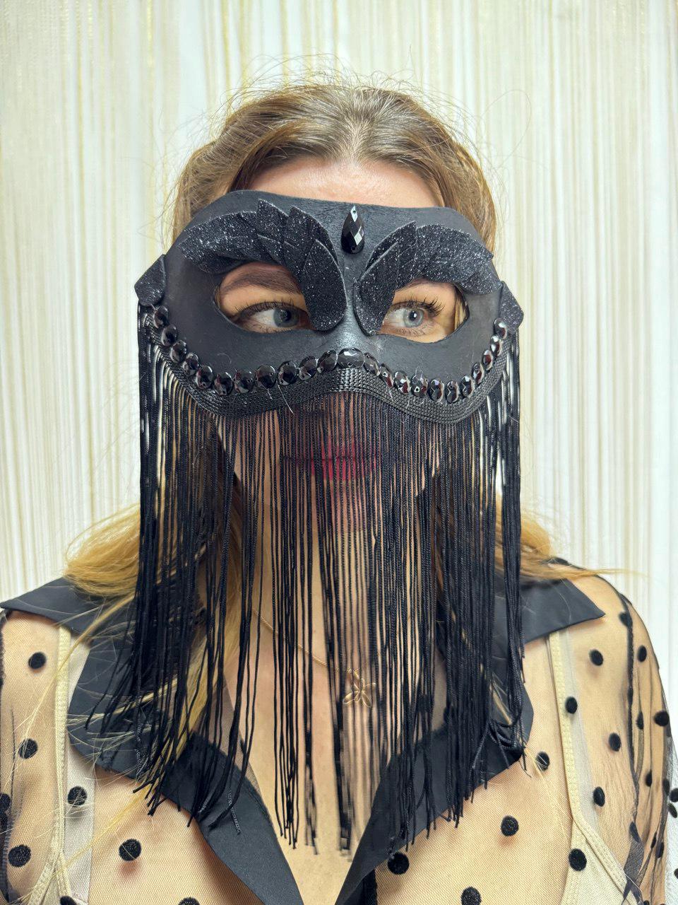 May include: Black masquerade mask with a decorative design. The mask features a beaded brow line, a central jewel, and fringe. The mask is made of a dark material and is worn over the eyes.