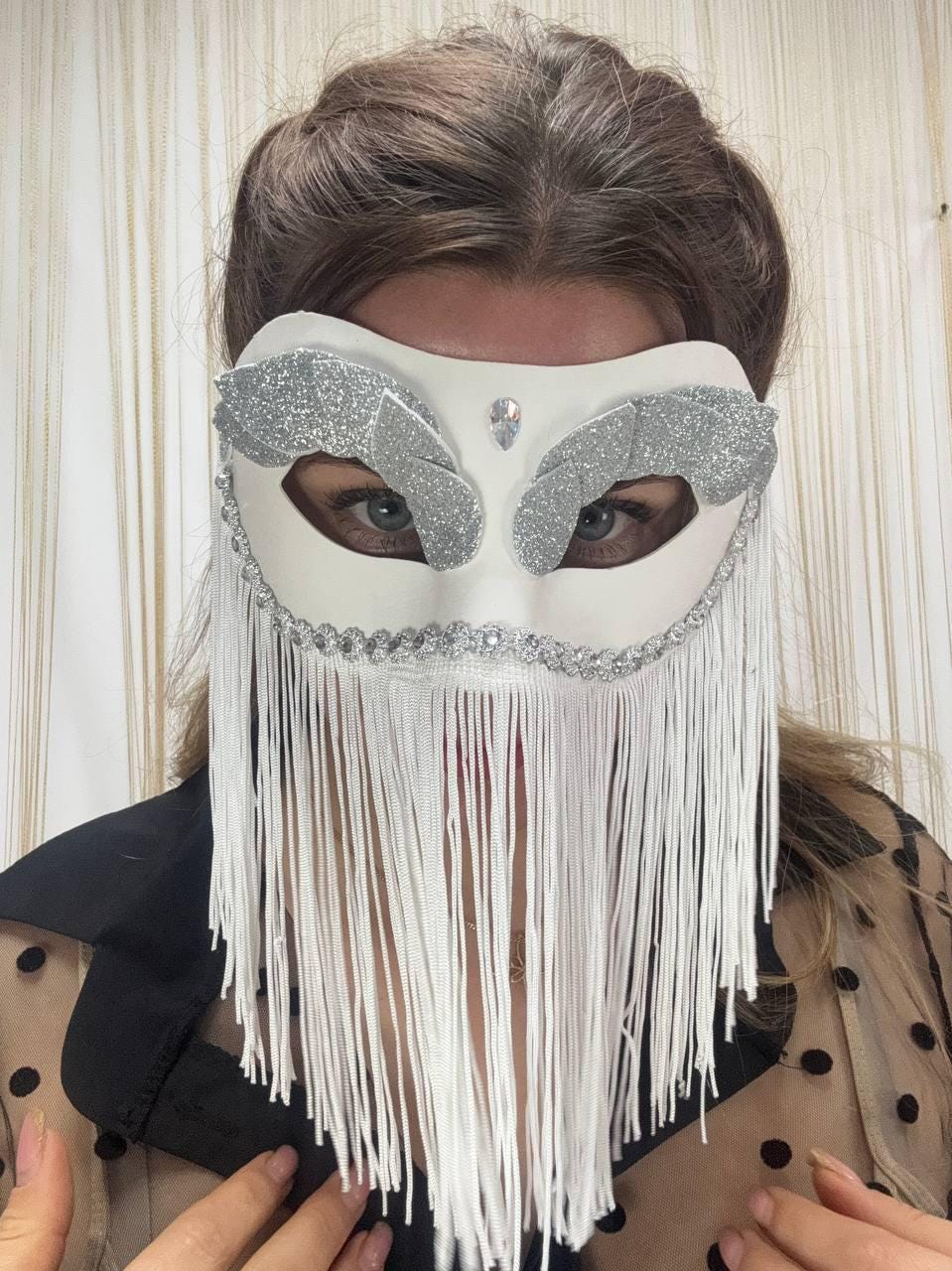 May include: White masquerade mask with silver glitter accents and a fringe. The mask features a central jewel and is decorated with silver rhinestones. The mask is designed for a costume or special event.