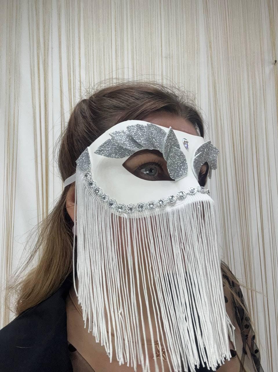 May include: White masquerade mask with silver glitter accents and a fringe of white strands. The mask features a decorative trim of small, clear rhinestones along the top edge. A white elastic band secures the mask.