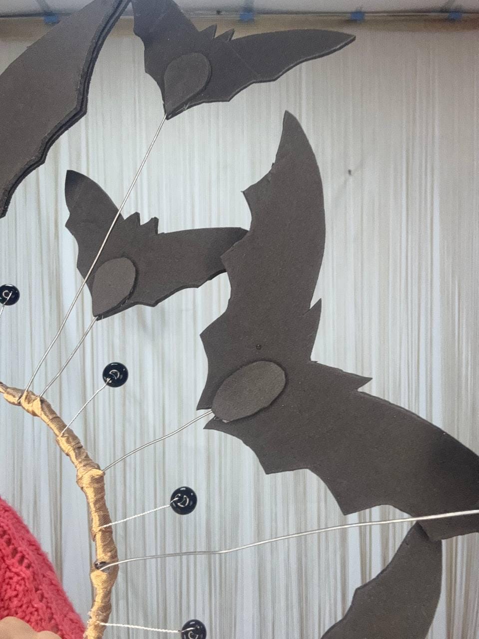 May include: A Halloween-themed decoration featuring several dark gray bat cutouts attached to thin wires, with a gold-colored branch-like structure. The bats have oval accents and are arranged against a white backdrop. Black beads are also attached.