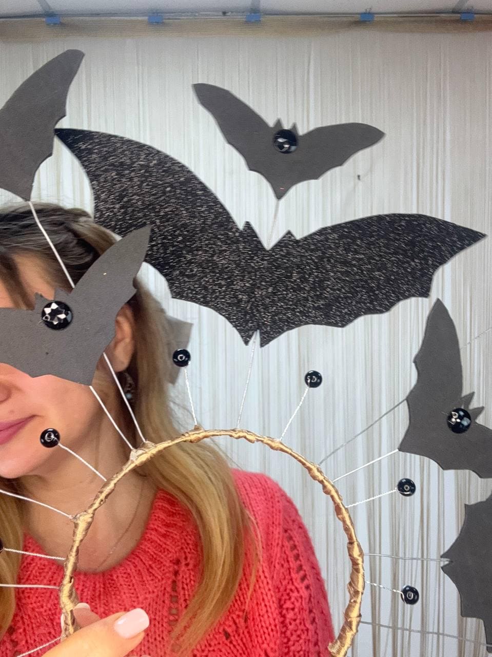May include: A Halloween-themed headpiece with a gold-colored circular frame and black bat decorations. The bats are made of black glitter paper and are attached to the frame with thin wires and black beads. The person in the image is wearing a red sweater.