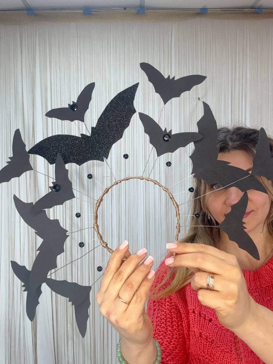May include: A Halloween-themed headpiece featuring a circular gold-toned wire frame adorned with black paper bats and black gem accents. The bats vary in size, with one made of glittery black material. A festive accessory for Halloween.