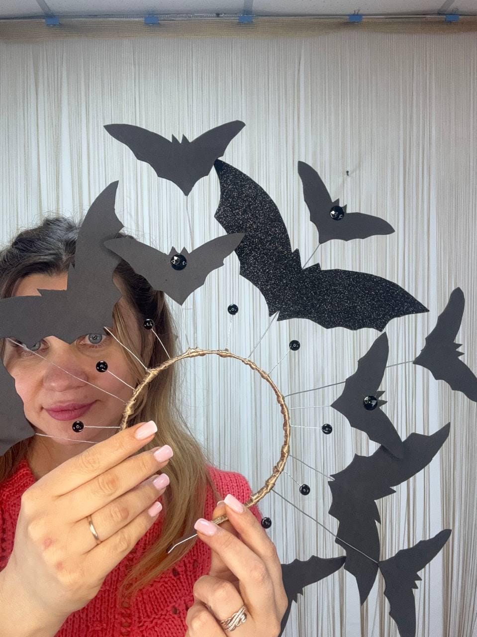 May include: A Halloween-themed headpiece featuring a gold-colored circular frame adorned with black bat cutouts. Some bats have glittered wings. Small black beads are attached to the frame and bats. The headpiece is designed for a festive occasion.