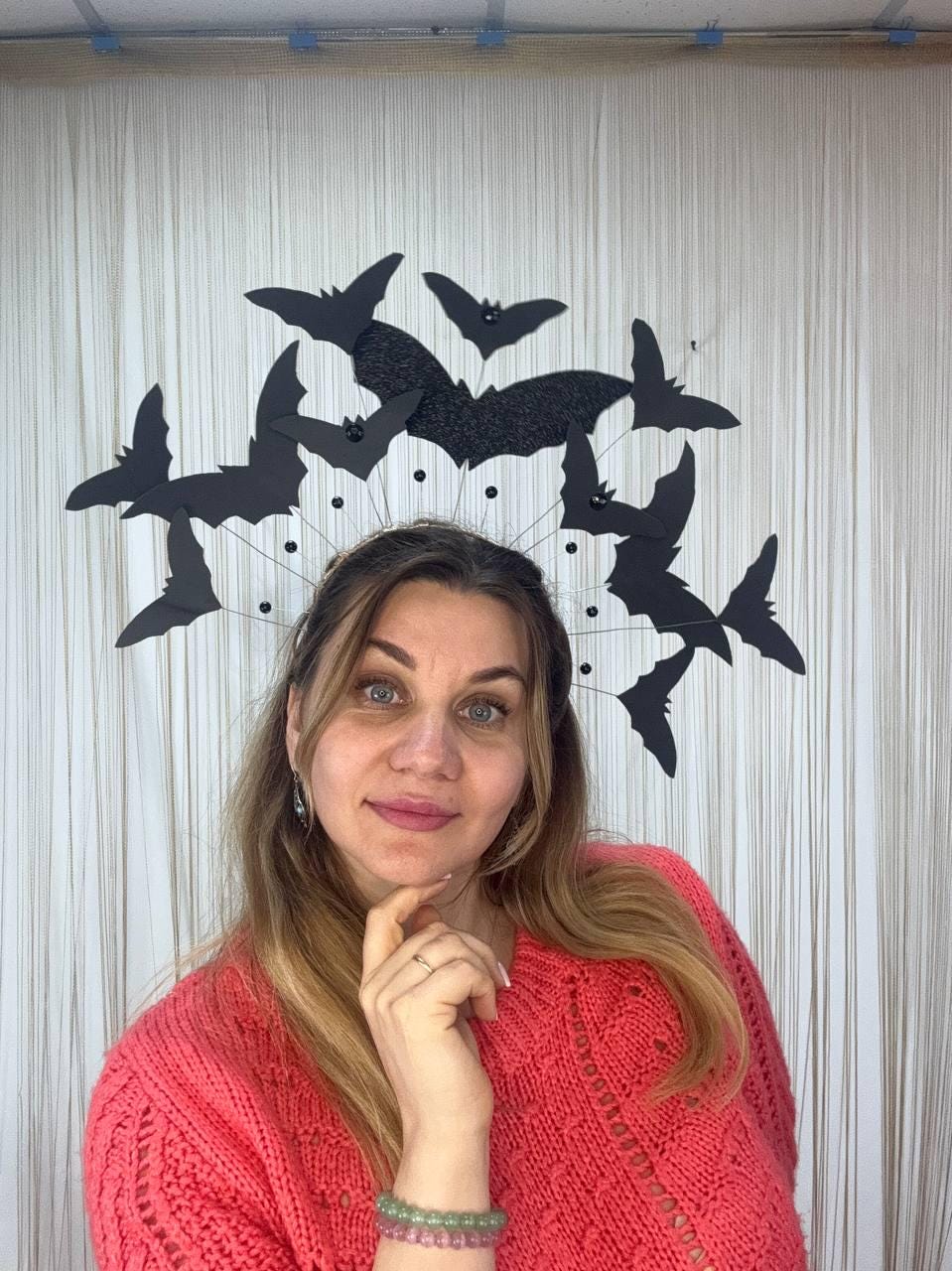 May include: A Halloween-themed headpiece featuring black bat silhouettes. The bats are arranged in a circular pattern, with a central glittery bat. The woman in the image is wearing a coral-colored sweater.