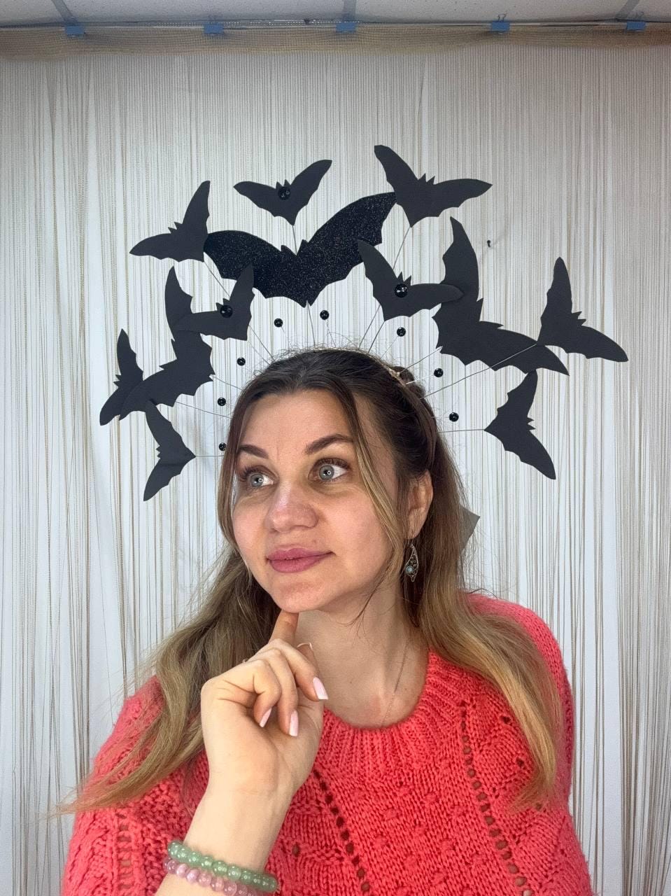 May include: A Halloween-themed headpiece featuring black bat silhouettes. The bats are arranged in a circular design, with a larger, glittery bat at the center. The headpiece is attached to a thin wire frame. The background is a white curtain.
