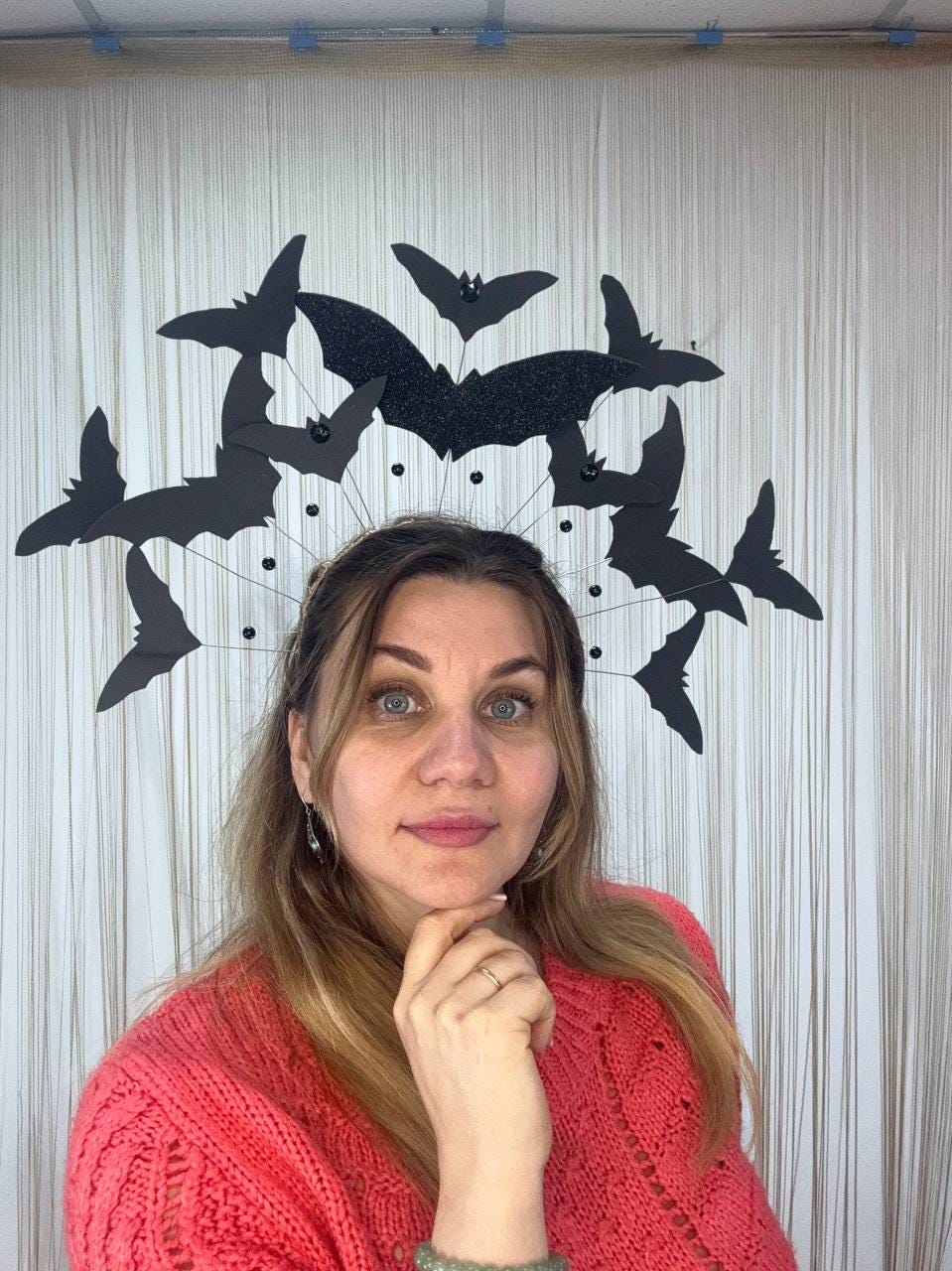 May include: A Halloween-themed headpiece featuring a halo of black bats. The bats are crafted from paper and arranged around a central, glittery bat. The headpiece is worn by a person wearing a coral-colored sweater.