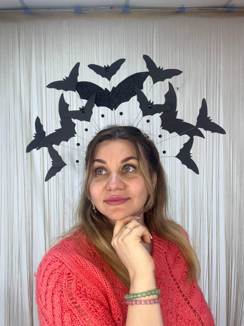 May include: A Halloween-themed headpiece featuring black bat silhouettes. The bats are arranged in a circular design, with a central glittery bat. The headpiece is worn by a person wearing a red sweater.
