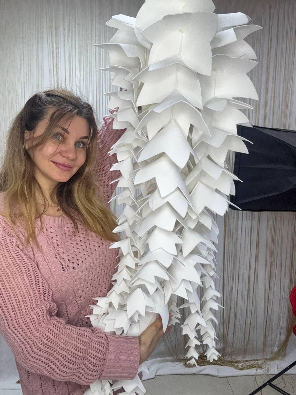 May include: A large, white, decorative hanging light fixture. The fixture is made of layered, pointed, leaf-like shapes. The light fixture is held by a person wearing a pink sweater.