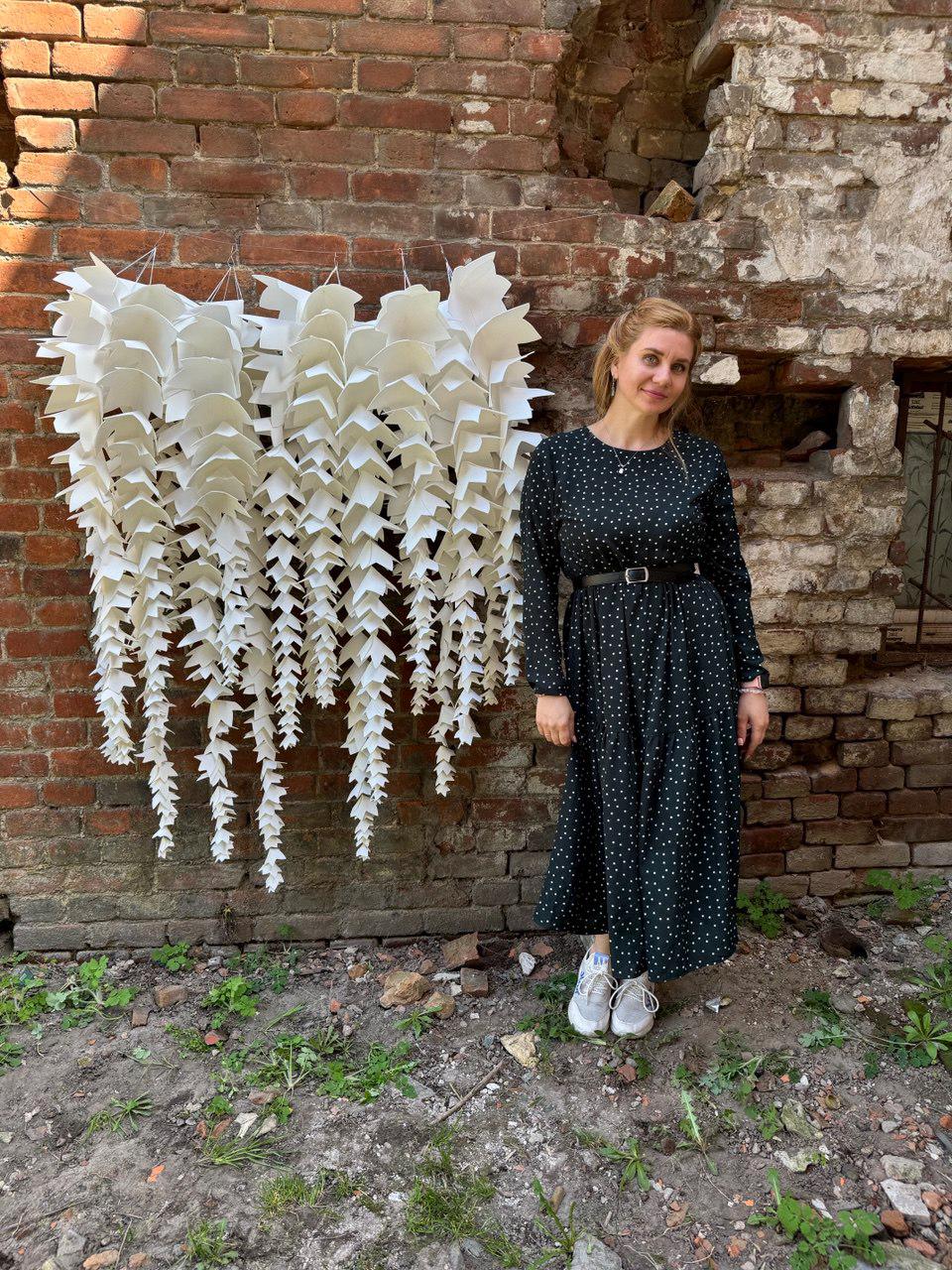 May include: A large, white, paper-like art installation hangs against a weathered brick wall. The piece is composed of numerous layered, leaf-shaped elements suspended from thin wires. A woman in a black polka-dot dress stands nearby.