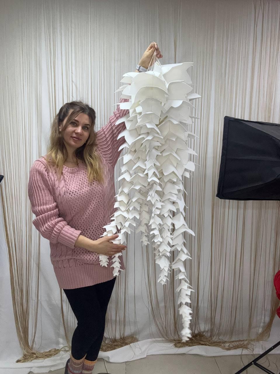 May include: A large, white paper sculpture with a cascading, layered design. The sculpture is held up by a hand and appears to be made of numerous folded paper elements, creating a textured, three-dimensional effect. The overall shape resembles a stylized plant or floral arrangement.