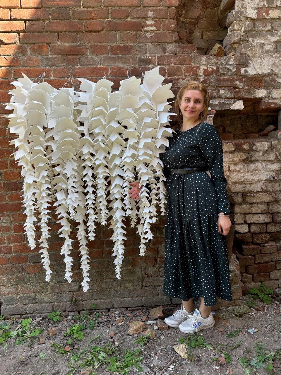 May include: A large, white, paper-like art installation with cascading, leaf-shaped elements hangs against a weathered brick wall. The artwork is the focal point, with a woman in a dark green polka-dot dress standing nearby.
