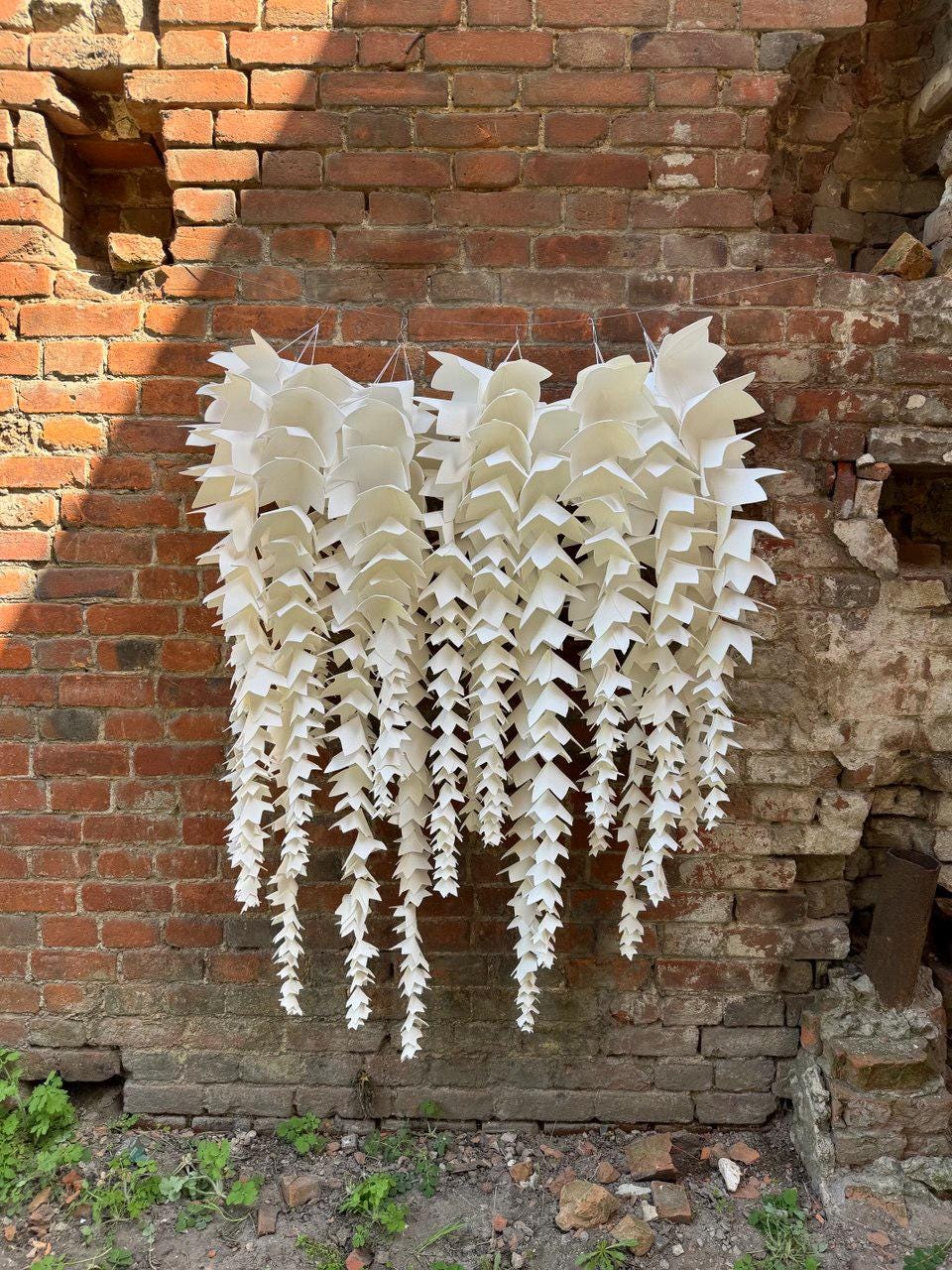 May include: A white, cascading paper art installation hangs against a weathered brick wall. The artwork features multiple strands of layered, geometric shapes, creating a textured, organic aesthetic. The piece is suspended by thin wires.