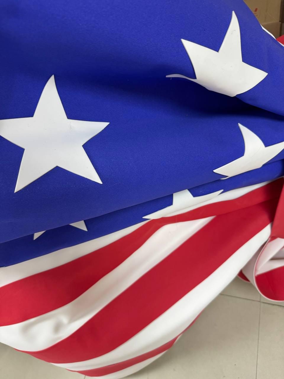 May include: A folded American flag with a blue field of stars and red and white stripes. The flag is made of a lightweight fabric and is partially folded, showing the stars and stripes.
